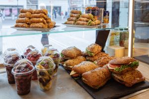 Eine Vitrine gefüllt mit verschiedenen belegten Brötchen, transparenten Bechern mit frischem Obst und aufgeschichteten Cannoli auf der oberen Ebene.