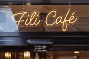 Ein leuchtender, warmweißer Neon-Schriftzug mit dem Namen „Fili Café“ hängt hinter einer Glasscheibe über dem Eingangsbereich.