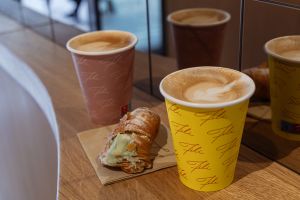 Zwei Coffee-to-go-Becher mit Latte Art in Hellbraun und Gelb stehen neben einem mit Pistaziencreme gefüllten, mit Puderzucker bestäubten Gebäck auf einer Holztheke.