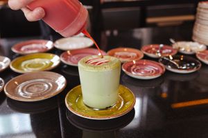 Eine Hand verziert einen grünen Matcha-Latte in Glas mit roter Soße