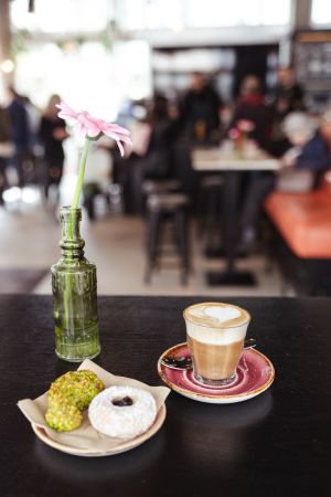 Ein Café-Tisch mit rosa Blume, Puderzucker-Donut, Pistaziengebäck und Kaffeegetränk