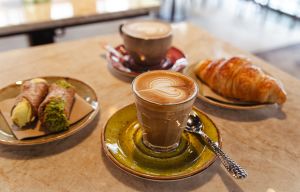 Kaffeetisch mit zwei Kaffegetränken, Croissant und zwei Pistazien-Gebäck-Teilchen.