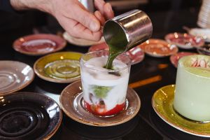 Eine Hand gießt grünen Matcha in ein Glas mit roter und weißer Flüssigkeit.