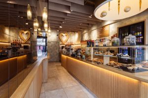 Café-Innenraum mit heller Holztheke, moderner Deckenbeleuchtung und einer großen Spiegelwand auf der linken Seite.