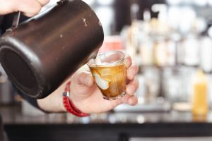 Barista gießt Milch aus Metallkännchen in ein kleines Kaffeeglas.