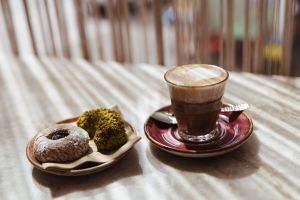 Kaffeeglas mit Milchschaum, daneben Teller mit Puderzucker-Donut und Pistaziengebäck auf hellem Tisch.